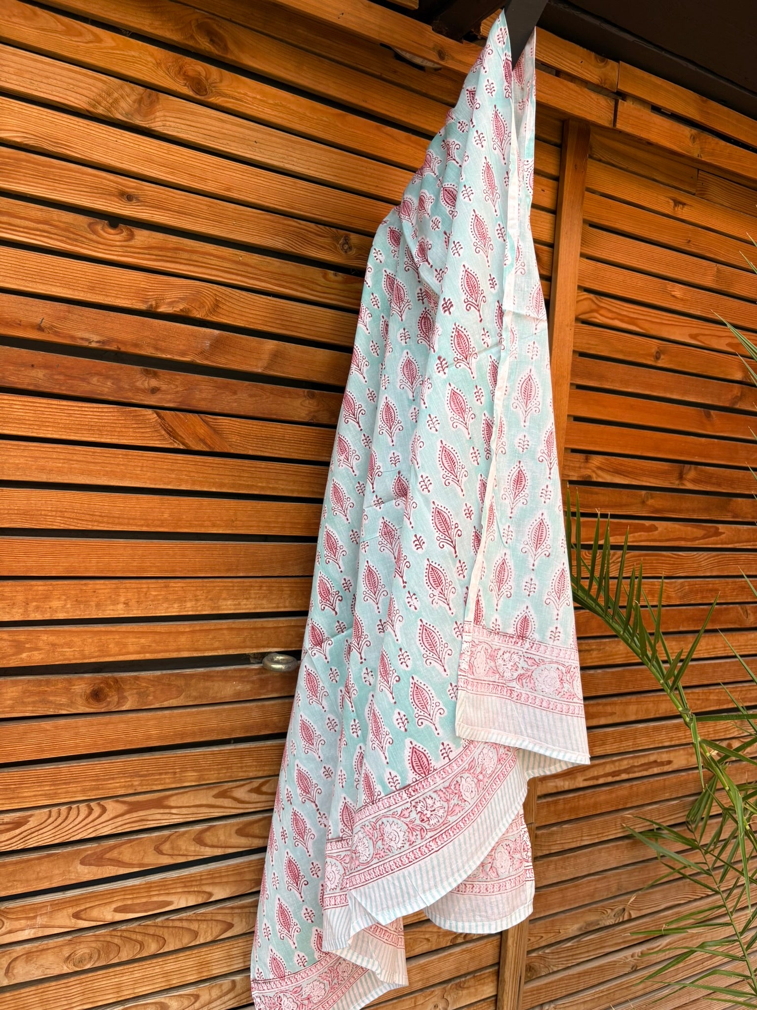 Paréo en coton léger avec impression artisanale indienne de type block print, présentant un motif répétitif de petits motifs floraux/botaniques de couleur rose fuchsia sur un fond vert menthe très clair. Une fine bordure ornementale assortie termine le tissu. Le paréo est suspendu naturellement devant un mur à claire-voie en bois clair et un feuillage vert.