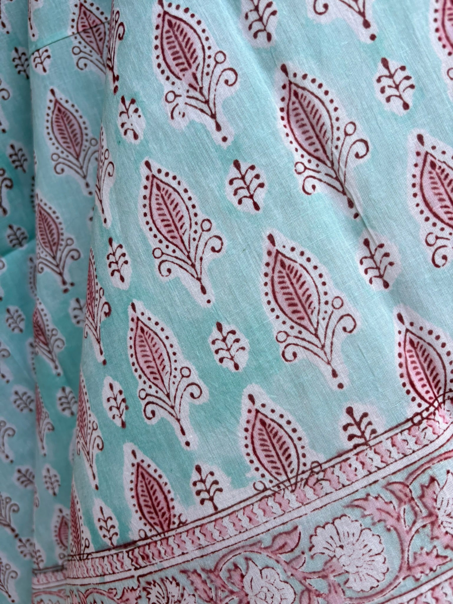 Paréo en coton léger avec impression artisanale indienne de type block print, présentant un motif répétitif de petits motifs floraux/botaniques de couleur rose fuchsia sur un fond vert menthe très clair. Une fine bordure ornementale assortie termine le tissu. 
