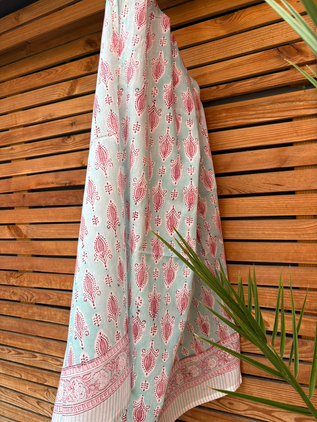 Paréo en coton léger avec impression artisanale indienne de type block print, présentant un motif répétitif de petits motifs floraux/botaniques de couleur rose fuchsia sur un fond vert menthe très clair. Une fine bordure ornementale assortie termine le tissu. Le paréo est suspendu naturellement devant un mur à claire-voie en bois clair et un feuillage vert.