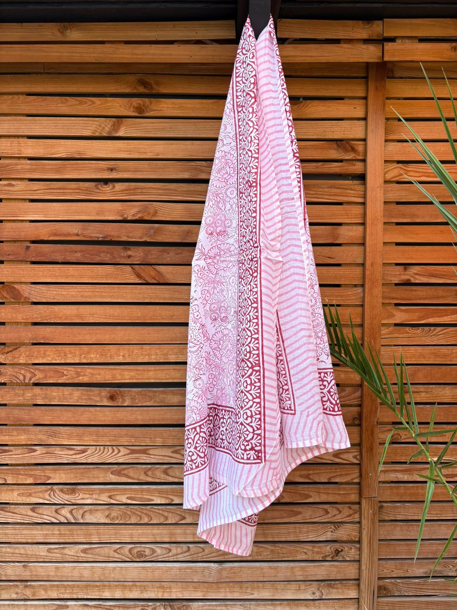 Paréo en coton léger avec impression artisanale indienne de type block print, présentant un motif floral complexe et une large bordure ornementale géométrique de couleur rouge écarlate sur fond blanc. Le tissu est suspendu naturellement devant un mur à claire-voie en bois clair et un feuillage vert
