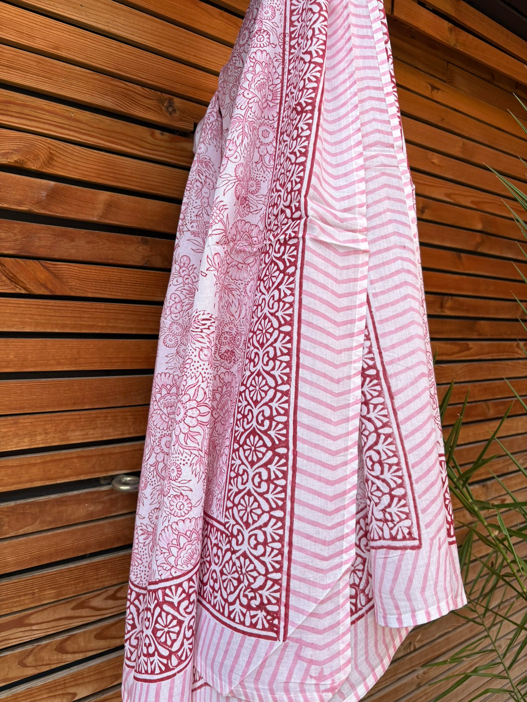 Paréo en coton léger avec impression artisanale indienne de type block print, présentant un motif floral complexe et une large bordure ornementale géométrique de couleur rouge écarlate sur fond blanc. Le tissu est suspendu naturellement devant un mur à claire-voie en bois clair et un feuillage vert