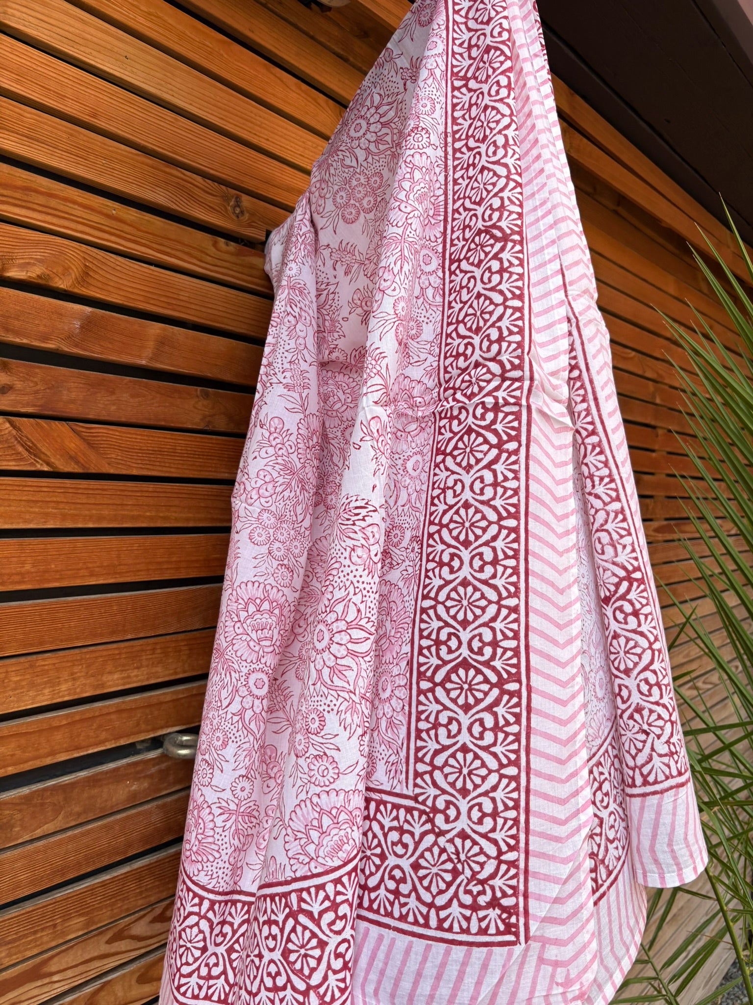 Paréo en coton léger avec impression artisanale indienne de type block print, présentant un motif floral complexe et une large bordure ornementale géométrique de couleur rouge écarlate sur fond blanc. Le tissu est suspendu naturellement devant un mur à claire-voie en bois clair et un feuillage vert