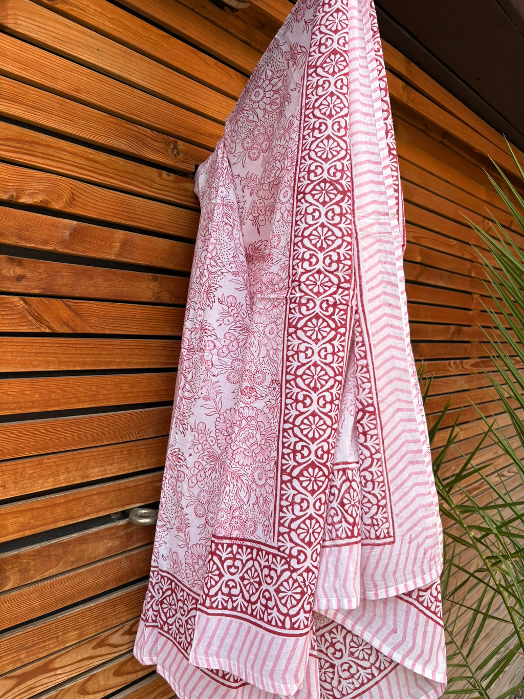 Paréo en coton léger avec impression artisanale indienne de type block print, présentant un motif floral complexe et une large bordure ornementale géométrique de couleur rouge écarlate sur fond blanc. Le tissu est suspendu naturellement devant un mur à claire-voie en bois clair et un feuillage vert