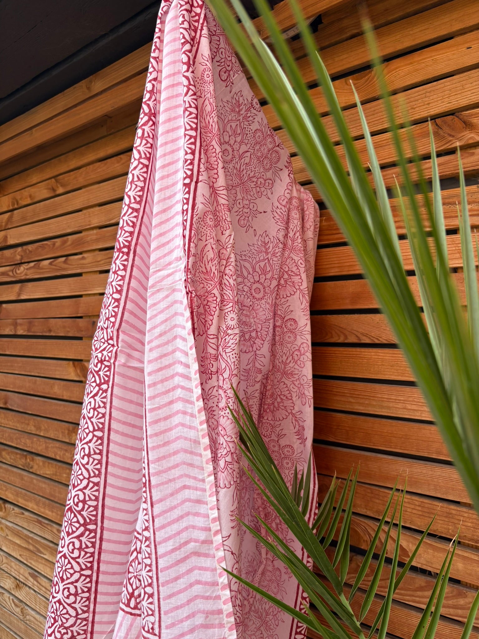 Paréo en coton léger avec impression artisanale indienne de type block print, présentant un motif floral complexe et une large bordure ornementale géométrique de couleur rouge écarlate sur fond blanc. Le tissu est suspendu naturellement devant un mur à claire-voie en bois clair et un feuillage vert