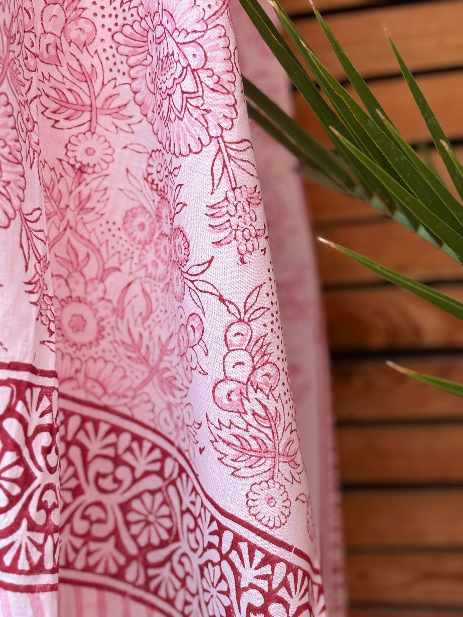 Paréo en coton léger avec impression artisanale indienne de type block print, présentant un motif floral complexe et une large bordure ornementale géométrique de couleur rouge écarlate sur fond blanc. Le tissu est suspendu naturellement devant un mur à claire-voie en bois clair et un feuillage vert
