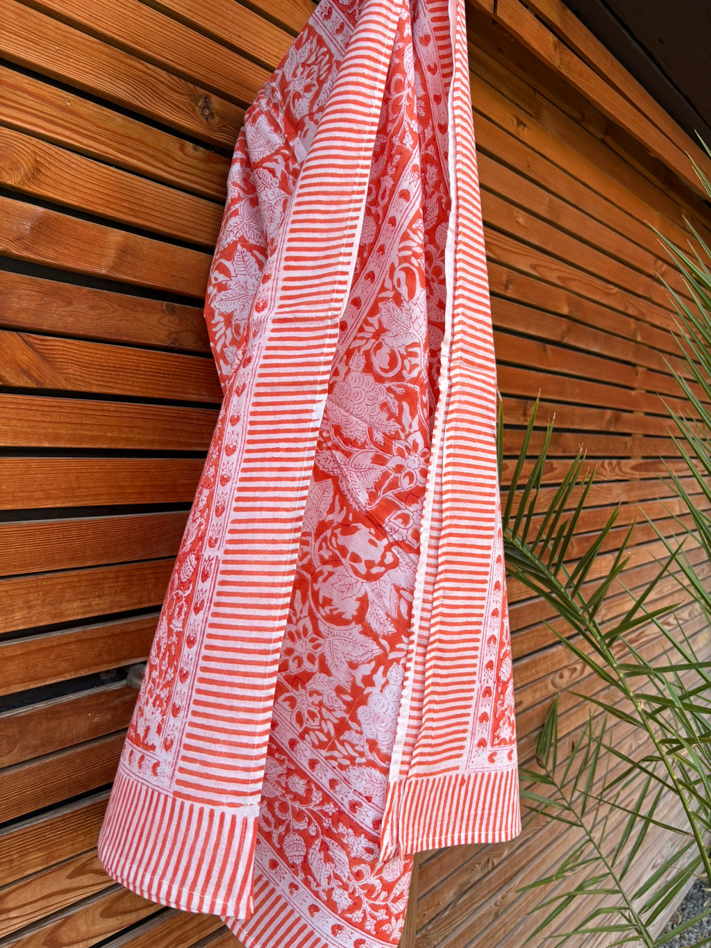 Paréo en coton léger avec impression artisanale indienne de type block print, présentant un motif floral complexe et une large bordure ornementale géométrique de couleur corail vif sur fond blanc. Le tissu est suspendu naturellement devant un mur à claire-voie en bois clair et un feuillage vert