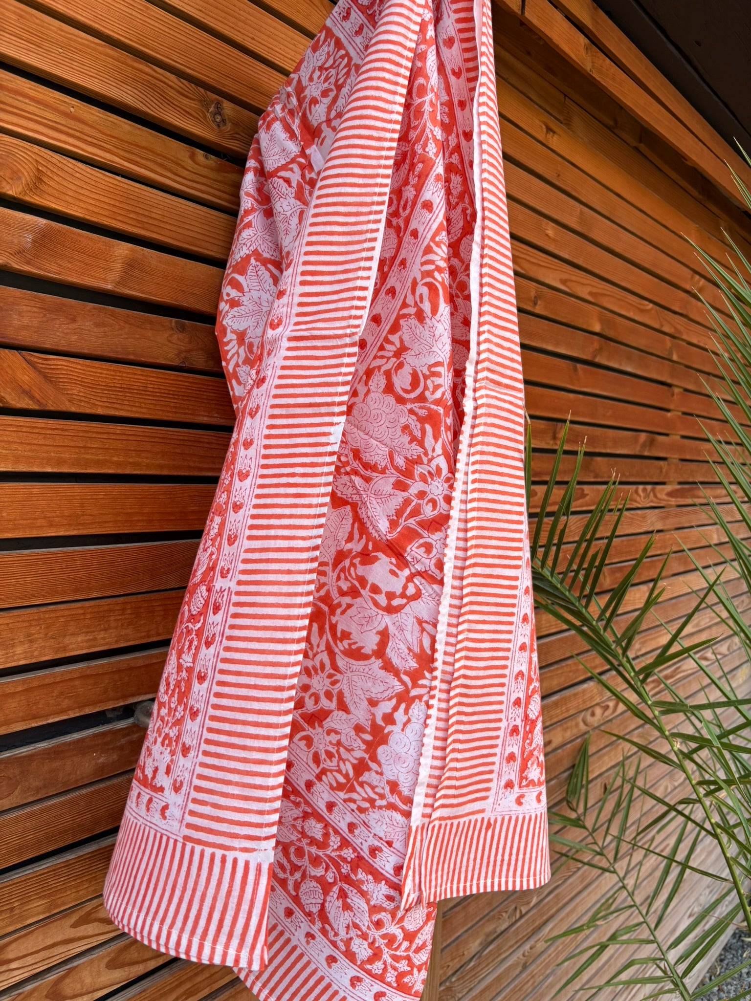 Paréo en coton léger avec impression artisanale indienne de type block print, présentant un motif floral complexe et une large bordure ornementale géométrique de couleur corail vif sur fond blanc. Le tissu est suspendu naturellement devant un mur à claire-voie en bois clair et un feuillage vert