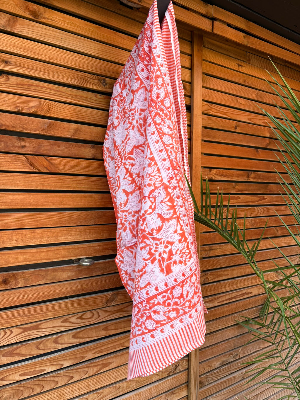 Paréo en coton léger avec impression artisanale indienne de type block print, présentant un motif floral complexe et une large bordure ornementale géométrique de couleur corail vif sur fond blanc. Le tissu est suspendu naturellement devant un mur à claire-voie en bois clair et un feuillage vert