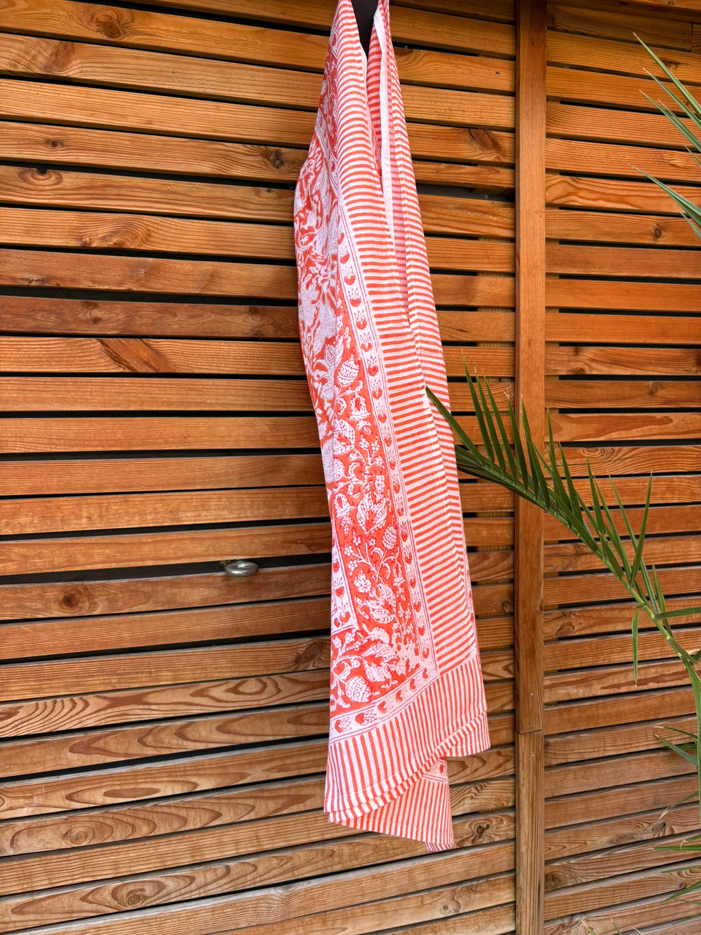 Paréo en coton léger avec impression artisanale indienne de type block print, présentant un motif floral complexe et une large bordure ornementale géométrique de couleur corail vif sur fond blanc. Le tissu est suspendu naturellement devant un mur à claire-voie en bois clair et un feuillage vert