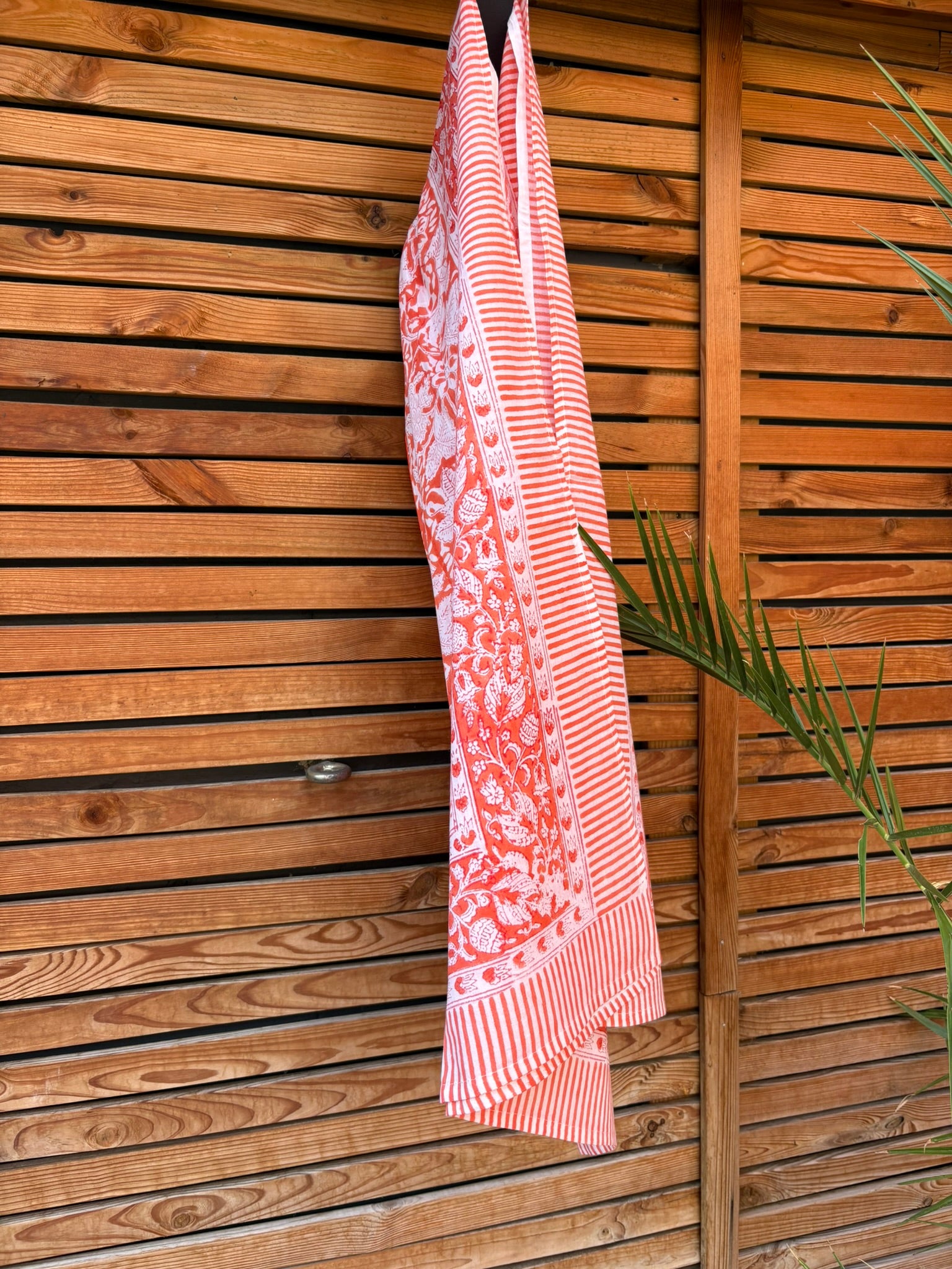 Paréo en coton léger avec impression artisanale indienne de type block print, présentant un motif floral complexe et une large bordure ornementale géométrique de couleur corail vif sur fond blanc. Le tissu est suspendu naturellement devant un mur à claire-voie en bois clair et un feuillage vert