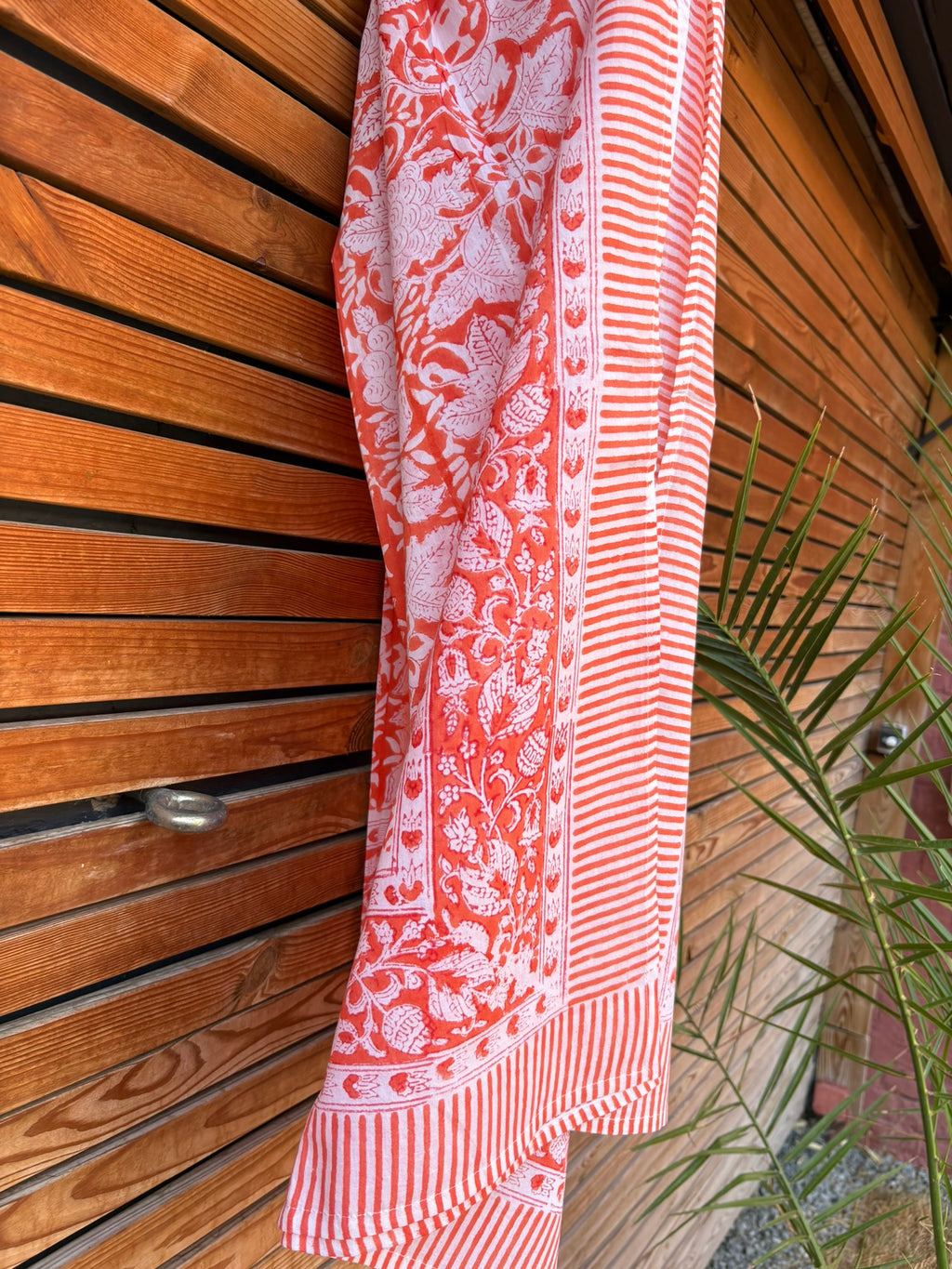Paréo en coton léger avec impression artisanale indienne de type block print, présentant un motif floral complexe et une large bordure ornementale géométrique de couleur corail vif sur fond blanc. Le tissu est suspendu naturellement devant un mur à claire-voie en bois clair et un feuillage vert