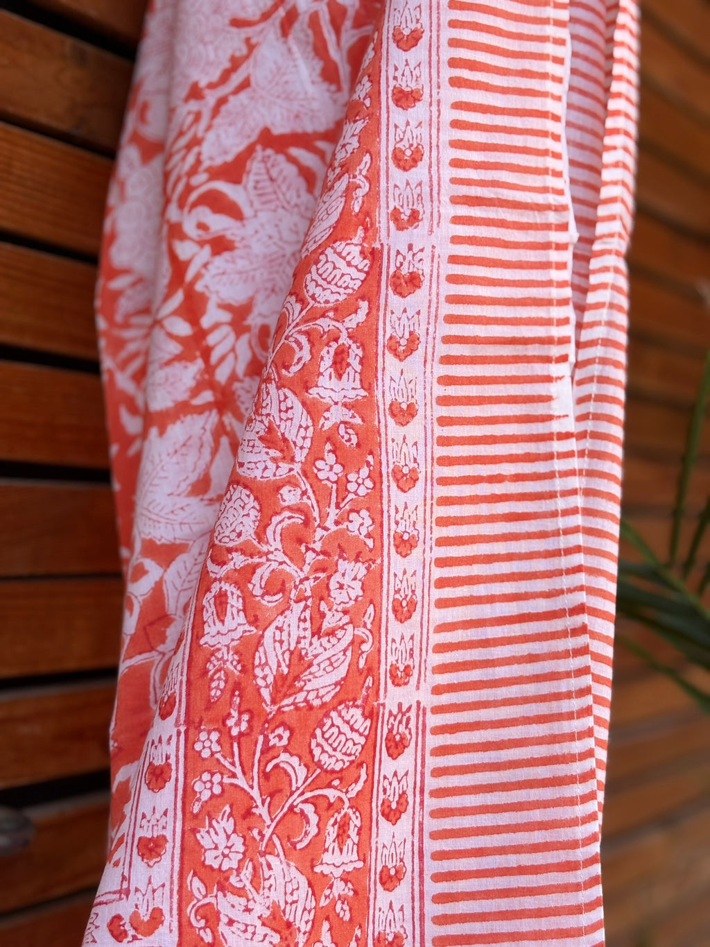 Paréo en coton léger avec impression artisanale indienne de type block print, présentant un motif floral complexe et une large bordure ornementale géométrique de couleur corail vif sur fond blanc. Le tissu est suspendu naturellement devant un mur à claire-voie en bois clair et un feuillage vert