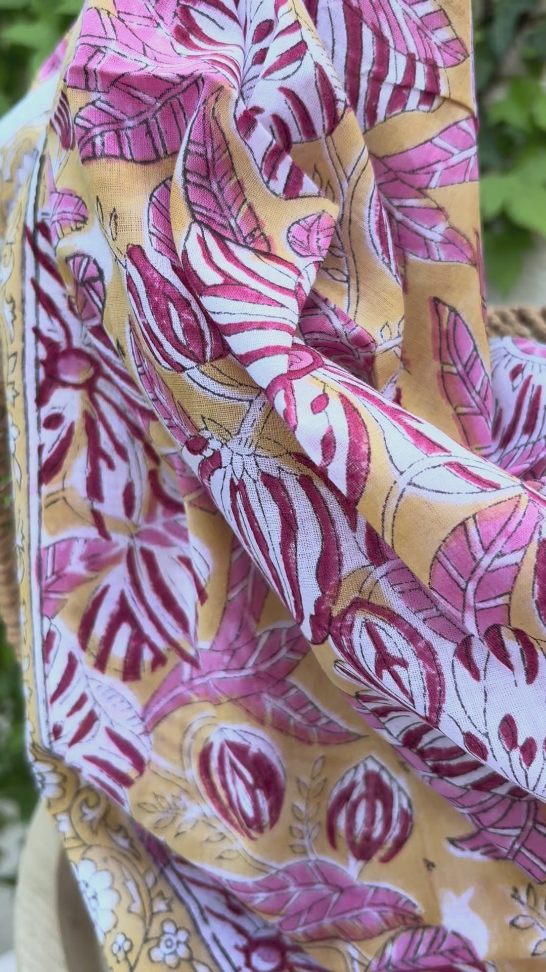 Un foulard en coton léger, imprimé selon la technique artisanale du blockprint, est présenté drapé sur une chaise en osier. Le design se compose de grandes fleurs épanouies et de feuilles striées dans des tons de rose framboise, bordeaux et blanc, le tout sur un fond jaune orangé chaleureux. Une bordure élégante sur fond blanc avec des motifs floraux noirs et jaunes vient terminer la pièce. L'arrière-plan, composé d'un mur clair et de lierre verdoyant, souligne l'aspect solaire et artisanal de l'accessoire.