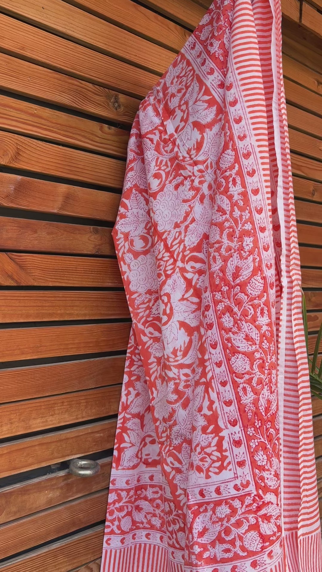 Paréo en coton léger avec impression artisanale indienne de type block print, présentant un motif floral complexe et une large bordure ornementale géométrique de couleur corail vif sur fond blanc. Le tissu est suspendu naturellement devant un mur à claire-voie en bois clair et un feuillage vert