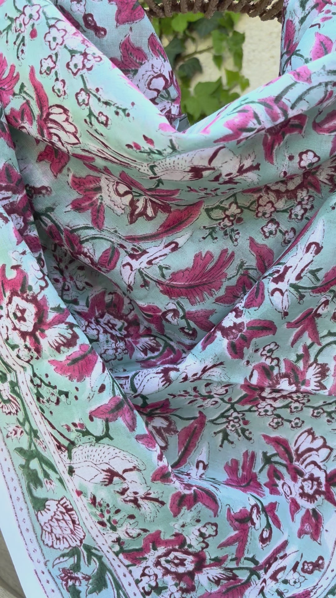 Un foulard en coton fin, réalisé avec la technique traditionnelle du blockprint, est présenté drapé sur une chaise en osier circulaire. Le motif se déploie sur un fond vert d'eau (céladon) et se compose de fleurs et de feuillages denses dans des tons de rose magenta, bordeaux et vert sapin. Une bordure blanche nette entoure le design floral. La mise en scène en extérieur, avec un mur clair et du lierre grimpant en arrière-plan, met en valeur les couleurs fraîches et le style bohème de l'accessoire.