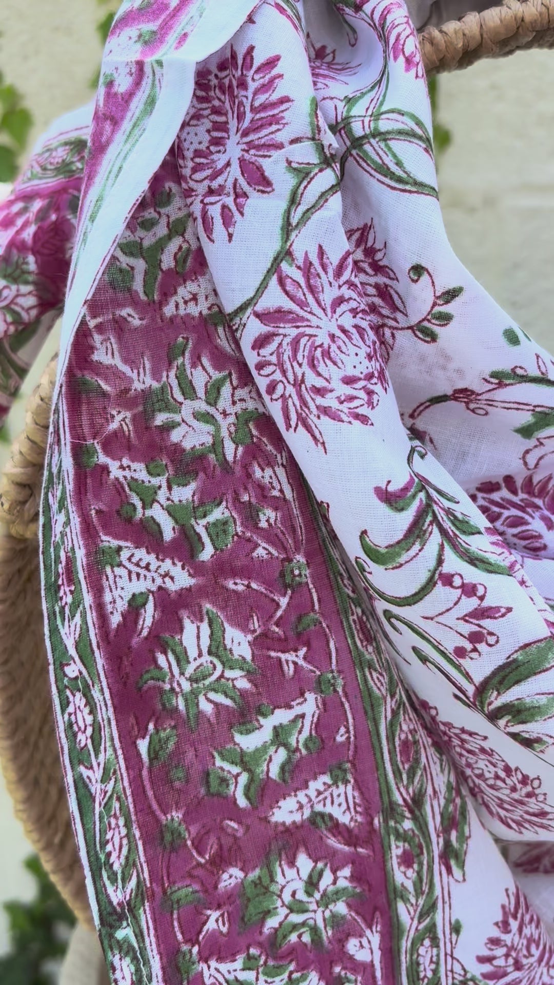 Un foulard en voile de coton blanc, léger et aérien, est présenté drapé sur une chaise en osier. Il est orné d'un imprimé artisanal blockprint représentant des fleurs de lotus et de chrysanthèmes stylisées dans des tons rose fuchsia et rose indien, reliées par des tiges et des feuilles vert tendre. Une large bordure fleurie assortie encadre la pièce. La mise en scène, avec un arrière-plan flou composé de lierre vert et d'un mur clair, souligne le caractère naturel, lumineux et bohème de cet accessoire